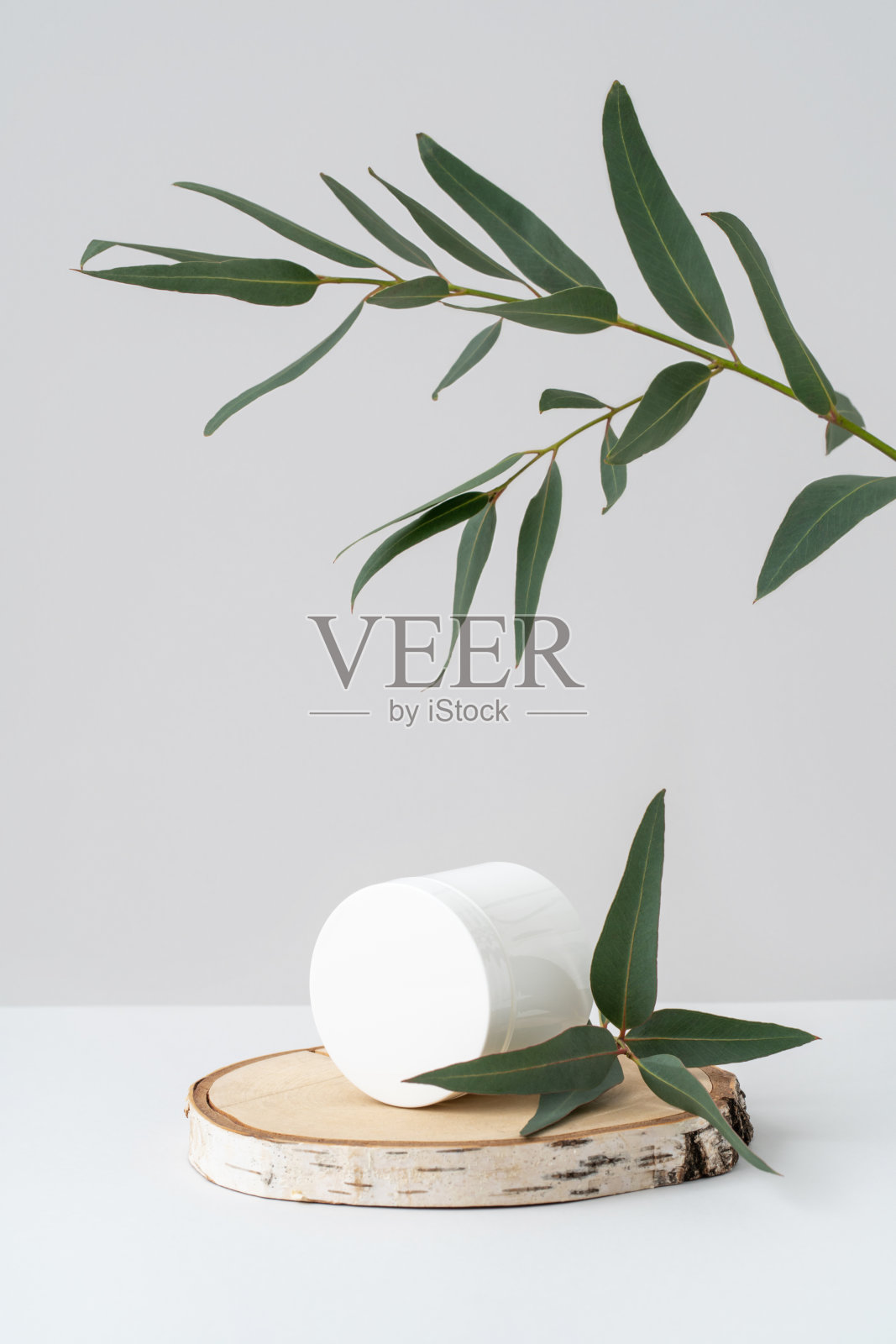 木质基座上的白色容器，灰色背景上的桉树枝。圆罐天然化妆品品牌桦木锯切和绿叶。照片摄影图片