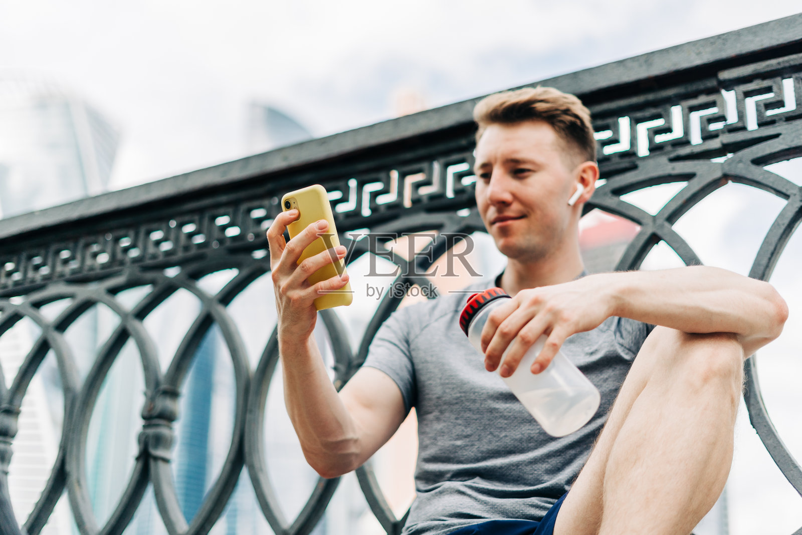 疲惫的年轻人坐在路上，使用手机和放松后的运动训练。在阴天的夏天，手持水瓶在城市街道上健身照片摄影图片