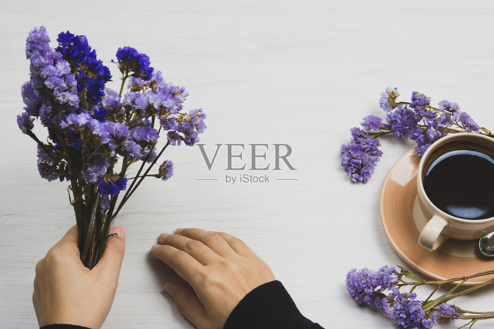 一个女人的手拿着一朵紫色的花，放在一张白色的木桌上，旁边是一杯咖啡。照片摄影图片