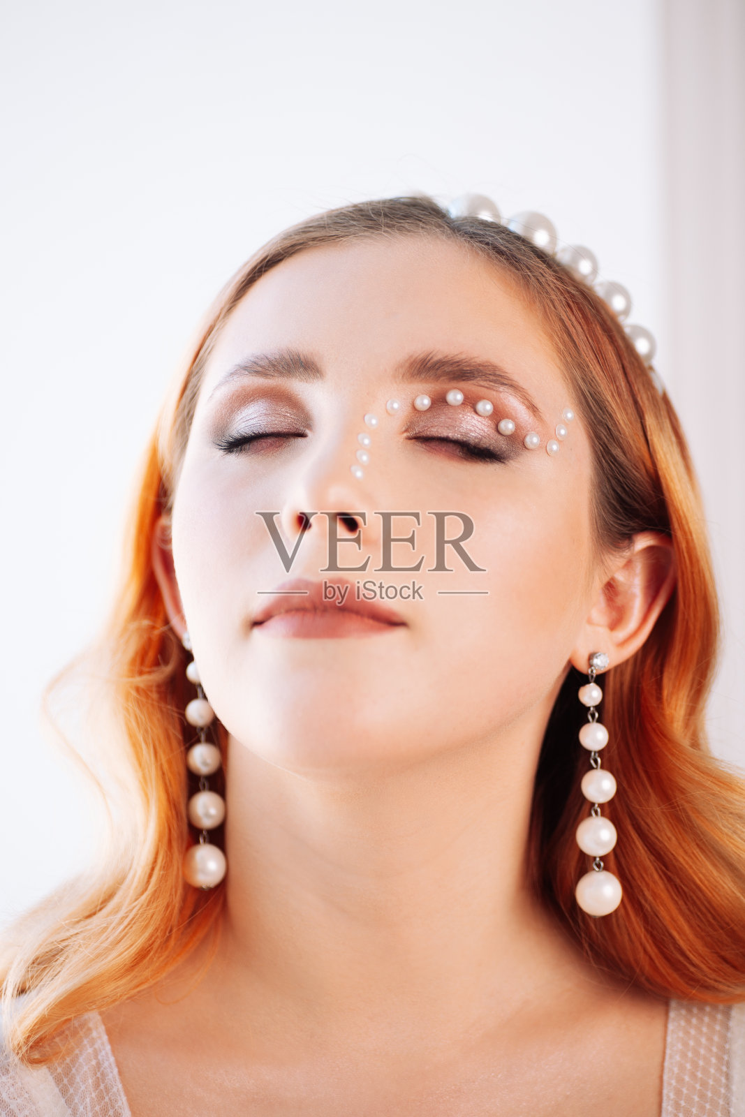 一个红色头发的女人闭着眼睛的垂直特写，用珍珠做的妆，珍珠边缘在她的头上和大耳环照片摄影图片