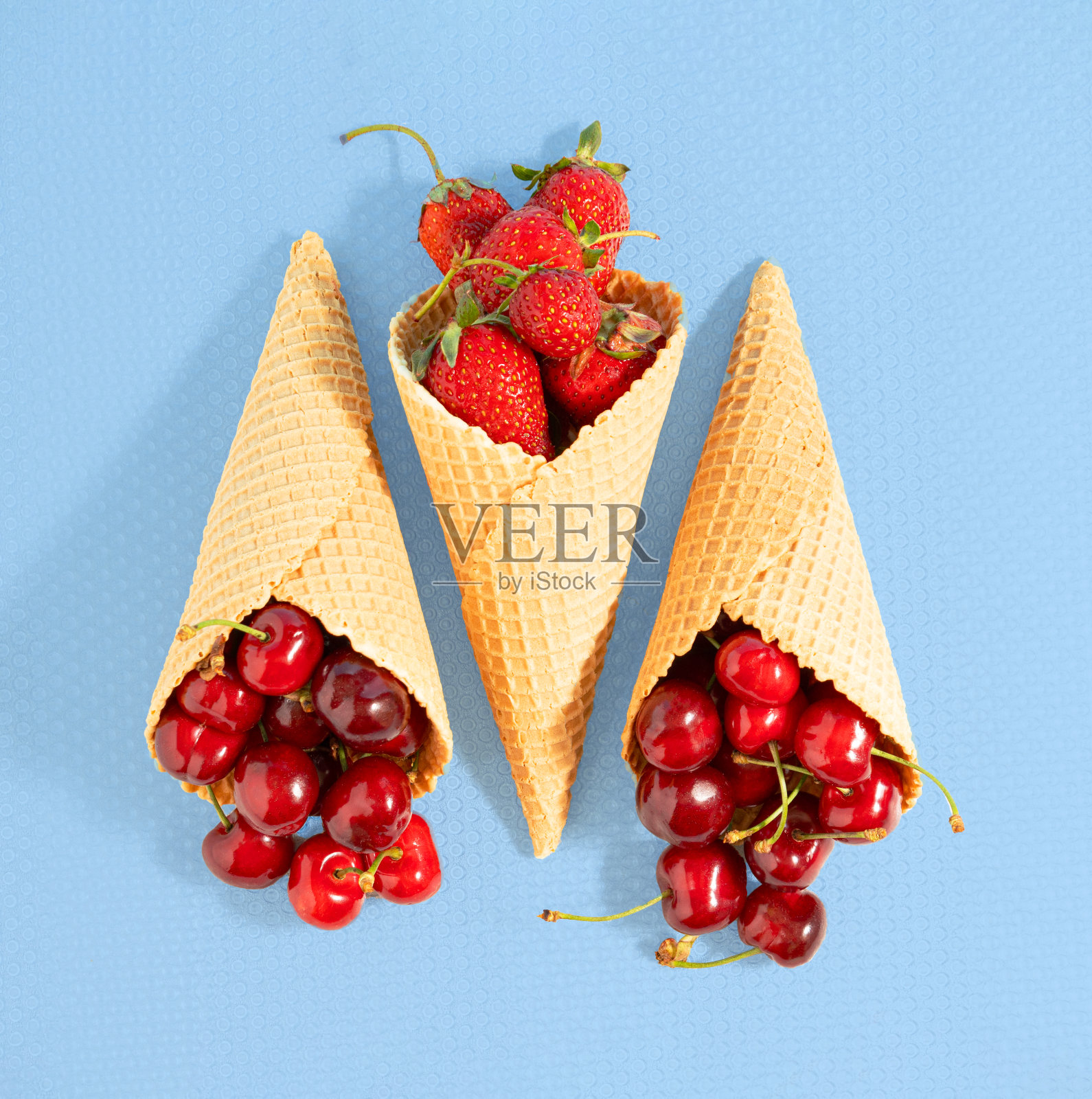 充满成熟草莓和樱桃的甜筒冰淇淋躺在蓝色的背景上。正方形平面敷设构成。简约夏日清凉概念照片摄影图片
