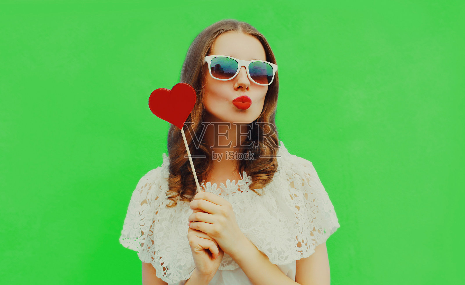 肖像近距离美丽的年轻女子吹她的嘴唇与红色的口红与甜蜜的心形棒棒糖在绿色的背景照片摄影图片