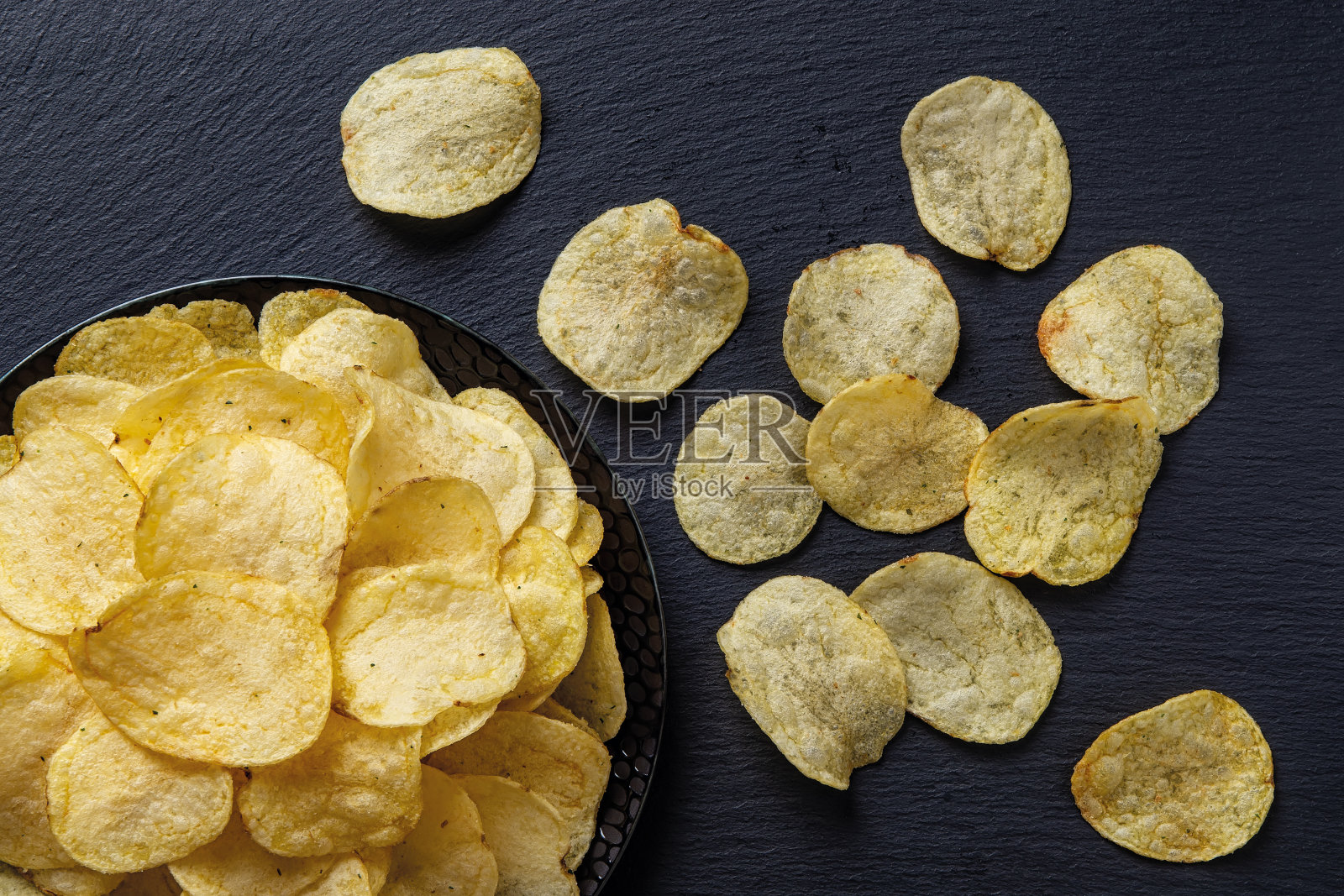 顶视图上的一个脆薯片上的黑色陶瓷盘子上的黑色石板表面。油炸土豆薄片的美味小吃。喜欢吃快餐、不健康的饮食和垃圾食品。照片摄影图片