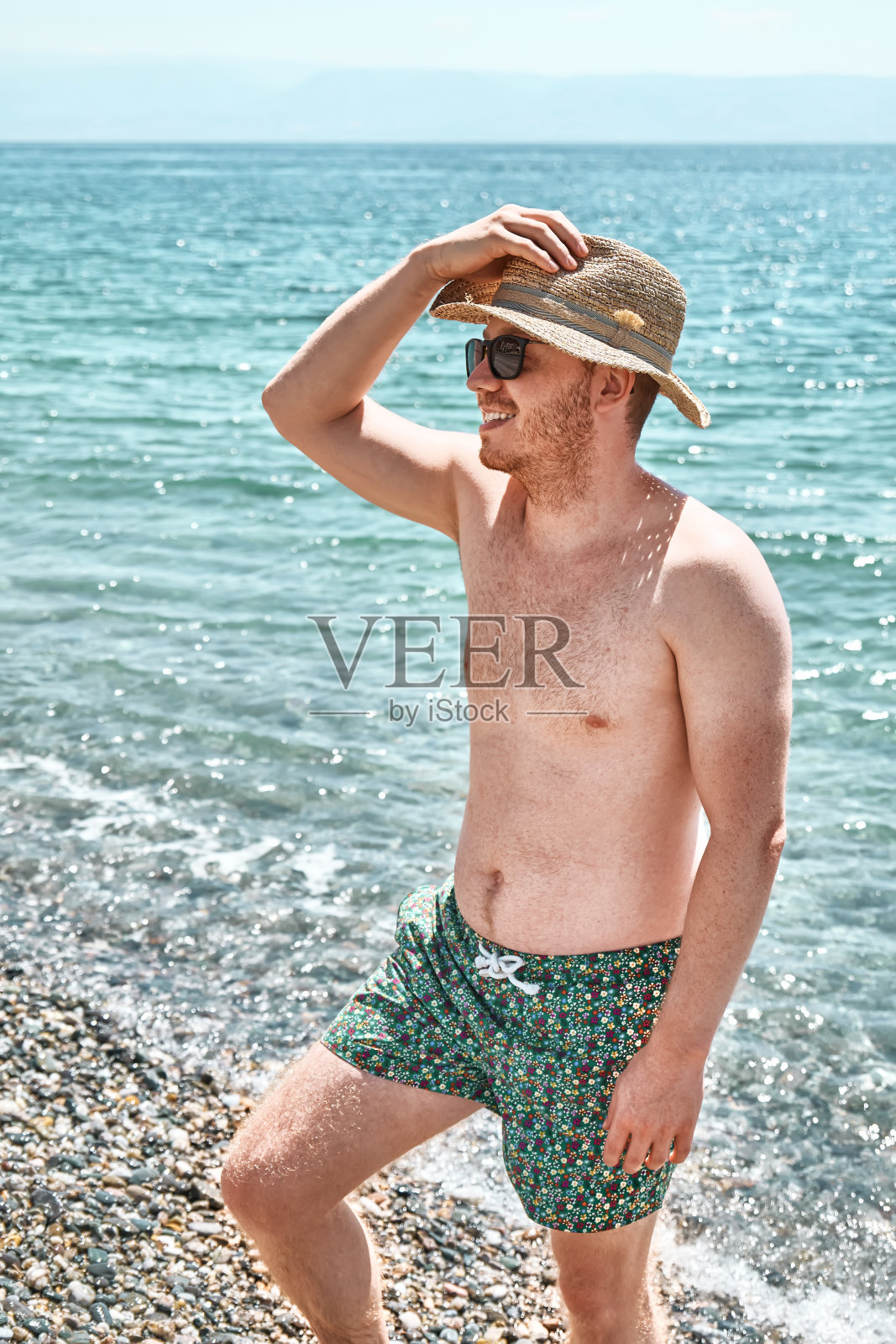 年轻英俊的红发男子赤膊站在沙滩上，穿着平角短裤，戴着太阳镜和草帽，露出肌肉健美的身材。一个在海滩上擦防晒油的男人。男人的护肤品。照片摄影图片