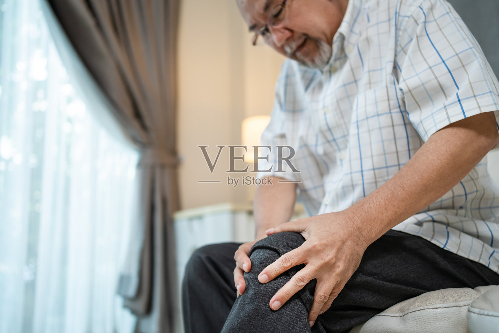 亚洲老人坐在养老院的沙发上，饱受腿伤之苦。痛苦的成熟老年人老年退休男性患者，患有膝关节问题。医院保险、医疗保健和医疗理念。照片摄影图片