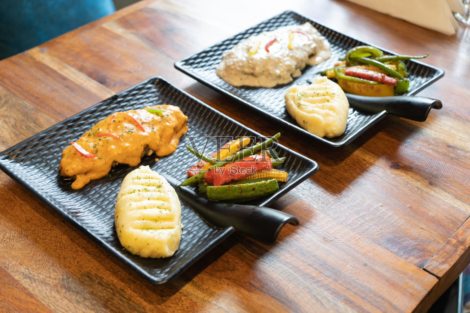 侧面的照片显示了两个美观的黑色带把手的方形盘子放在木桌上，里面装满了白酱意大利面和红酱土豆泥和辣椒蔬菜沙拉照片摄影图片