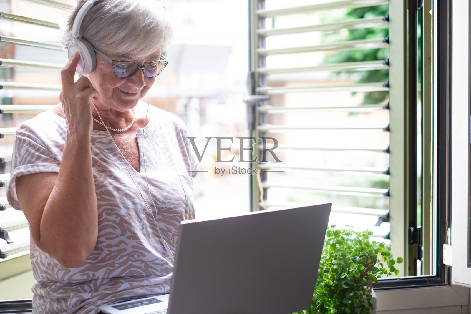 迷人的资深女子坐在靠近窗户使用笔记本电脑，戴着耳机。微笑的白人女性享受科技和社交媒体照片摄影图片