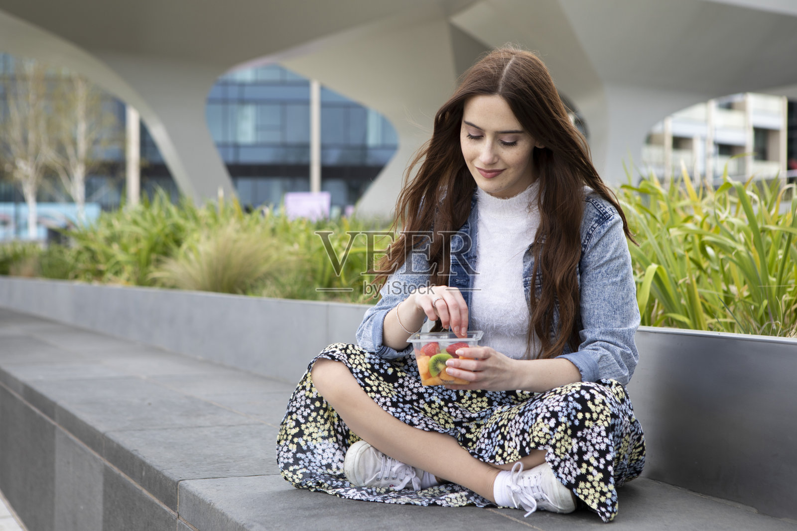 一名年轻女子穿着碎花裙、白色套头衫和牛仔夹克，盘腿坐在混凝土座椅上，吃着塑料(聚对苯二甲酸乙二醇酯)容器里的水果照片摄影图片