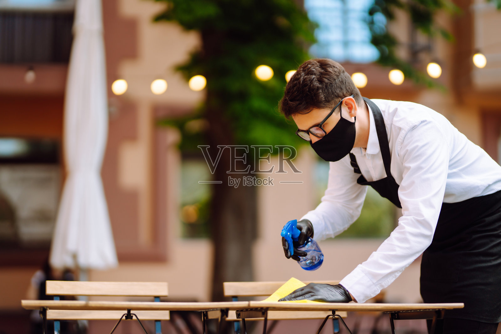 消毒预防COVID-19。检疫城市咖啡馆服务员戴着防护口罩和手套，用消毒剂喷雾和超细纤维布清洁桌子。照片摄影图片