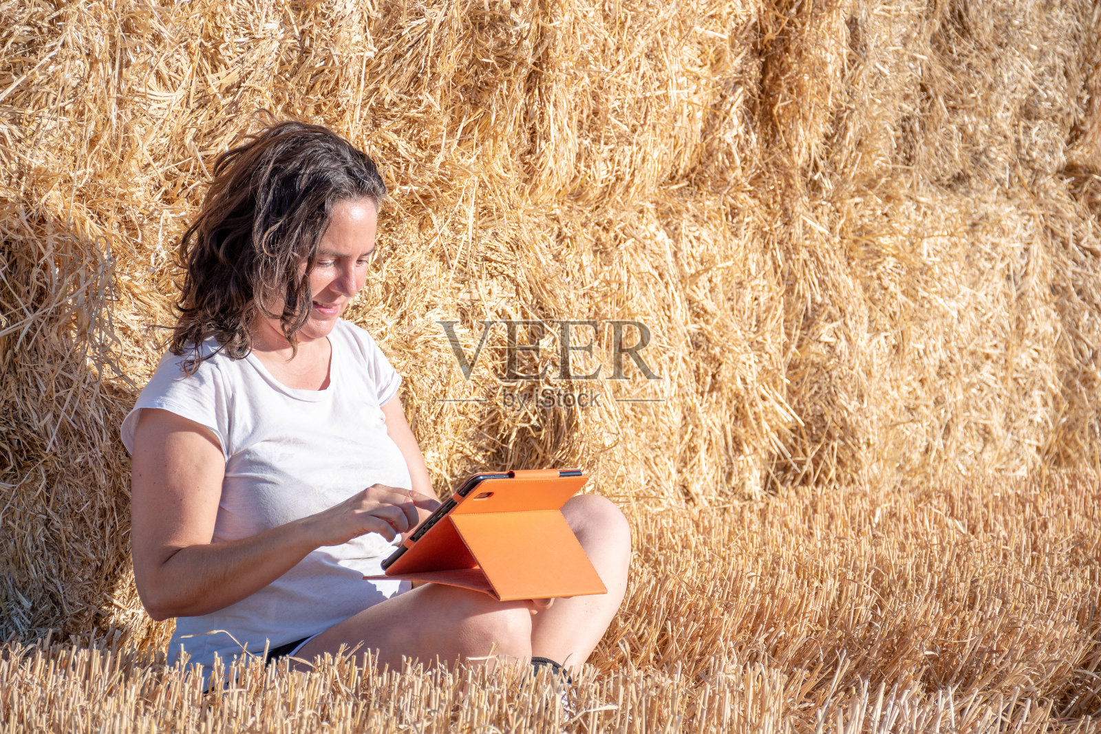 中年黑发的拉丁妇女坐在地上在一个领域靠在一堆稻草，使用一个平板与橙色覆盖照片摄影图片