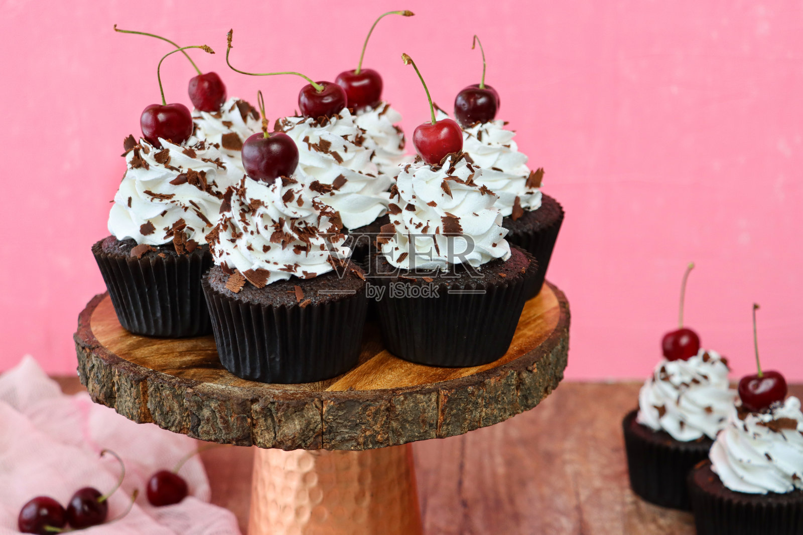 这是一批自制的黑森林蛋糕，在铜制和木质蛋糕架上的棕色纸制蛋糕盒里，插孔状的奶油玫瑰结上撒有巧克力屑的羊奶樱桃，粉红色的薄纱，粉色的背景，突出前景照片摄影图片