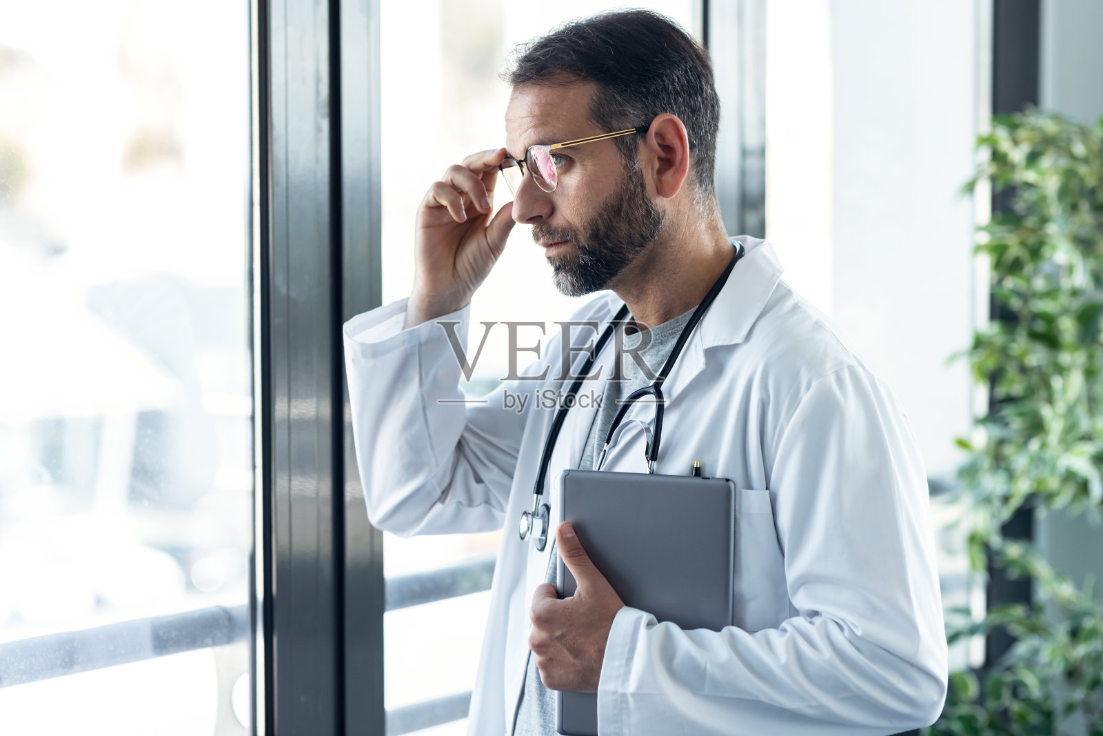 英俊成熟的男医生一边工作一边拿着平板电脑望着窗户，站在医务室里会诊。照片摄影图片