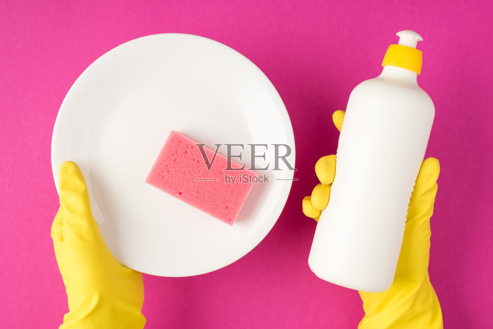 俯视图照片的手在黄色橡胶手套拿着白色洗涤剂瓶没有标签白色清洁板和粉红色洗涤垫孤立的粉红色背景与copyspace照片摄影图片