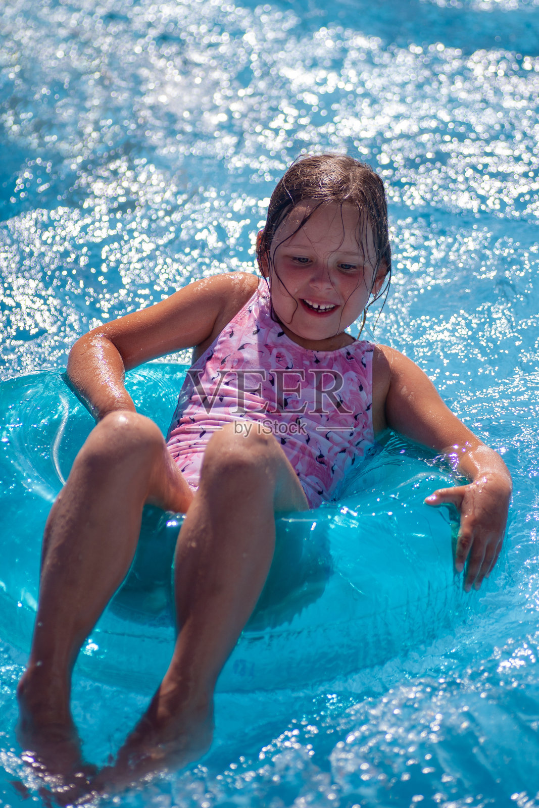 一个快乐的笑着的女孩穿着粉红色的泳衣，在阳光下闪闪发光的海面或泳池表面上骑着一个透明的充气圈照片摄影图片