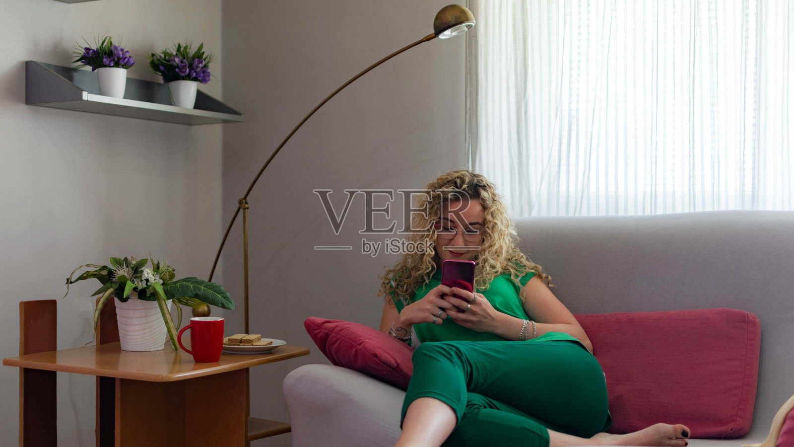 美丽的年轻女子卷发，坐在她的家，在她的阳台上，躺下，用她的智能手机与她的朋友联系，微笑着享受她的空闲时间，放松的咖啡和饼干。照片摄影图片