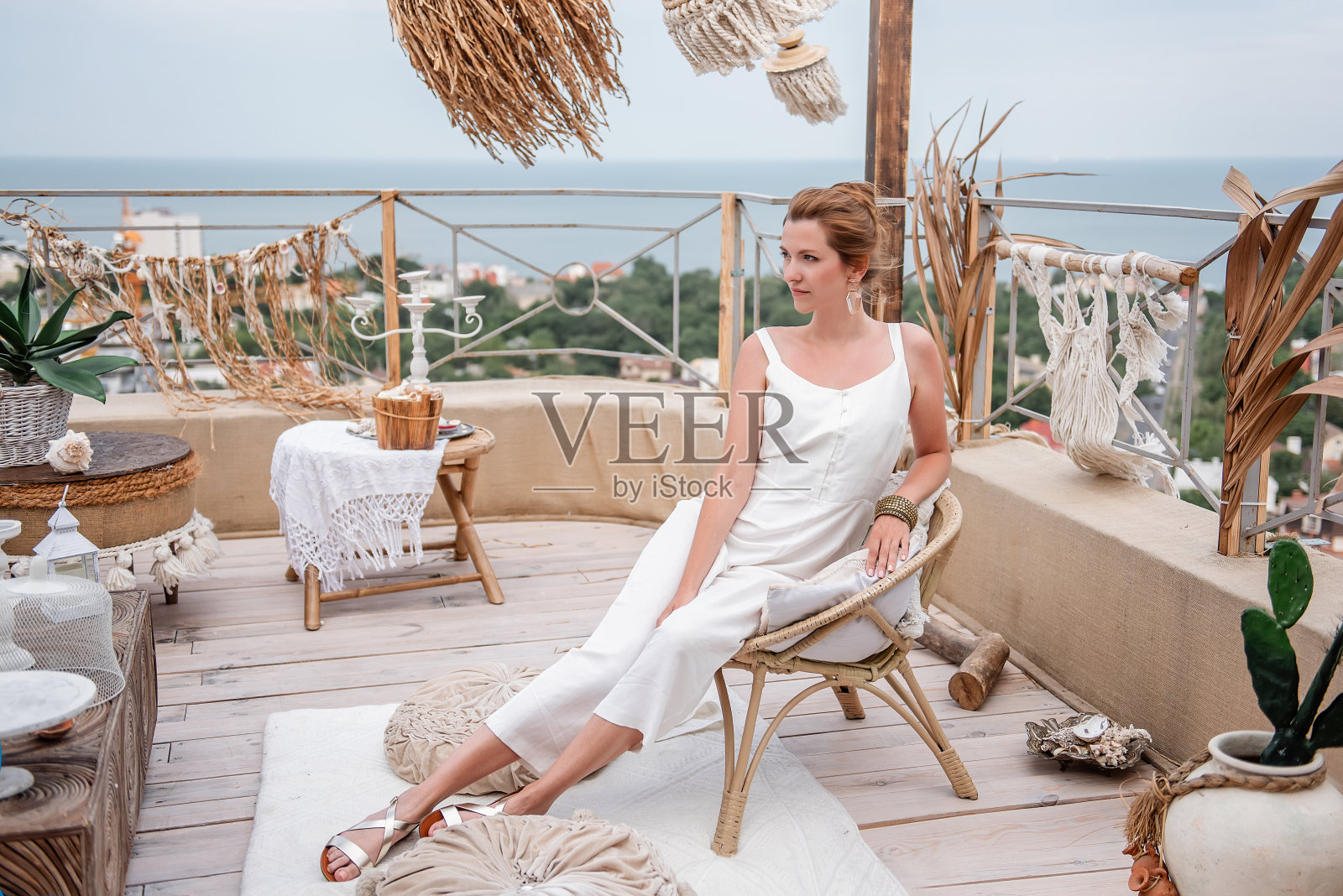 一名年轻女子穿着时尚的白色连身衣，坐在户外屋顶露台上的复古藤椅上，俯瞰着大海。印度风格的室内，摩洛哥沙色调，流苏，潘帕斯草照片摄影图片