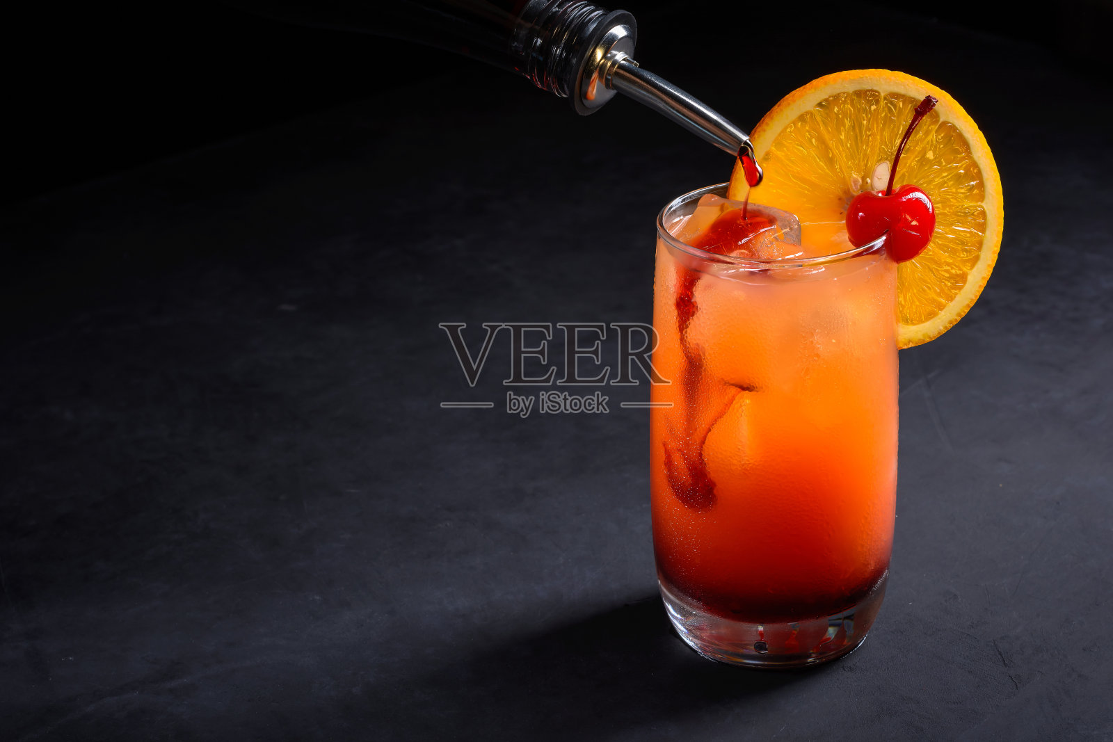制作龙舌兰日出鸡尾酒。格林纳丁慢慢地倒入一杯冰、橙汁和龙舌兰酒。深色的石头背景与复制空间。照片摄影图片