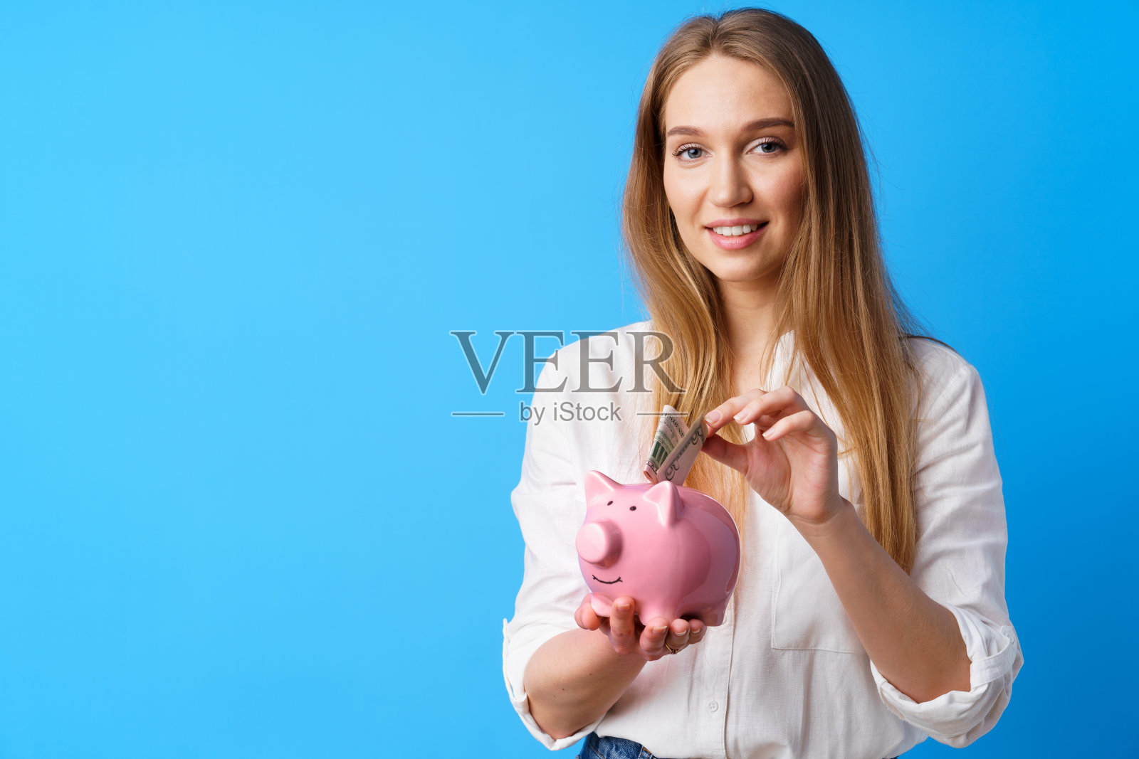 美丽微笑的年轻女子与小猪储蓄罐在蓝色的背景照片摄影图片