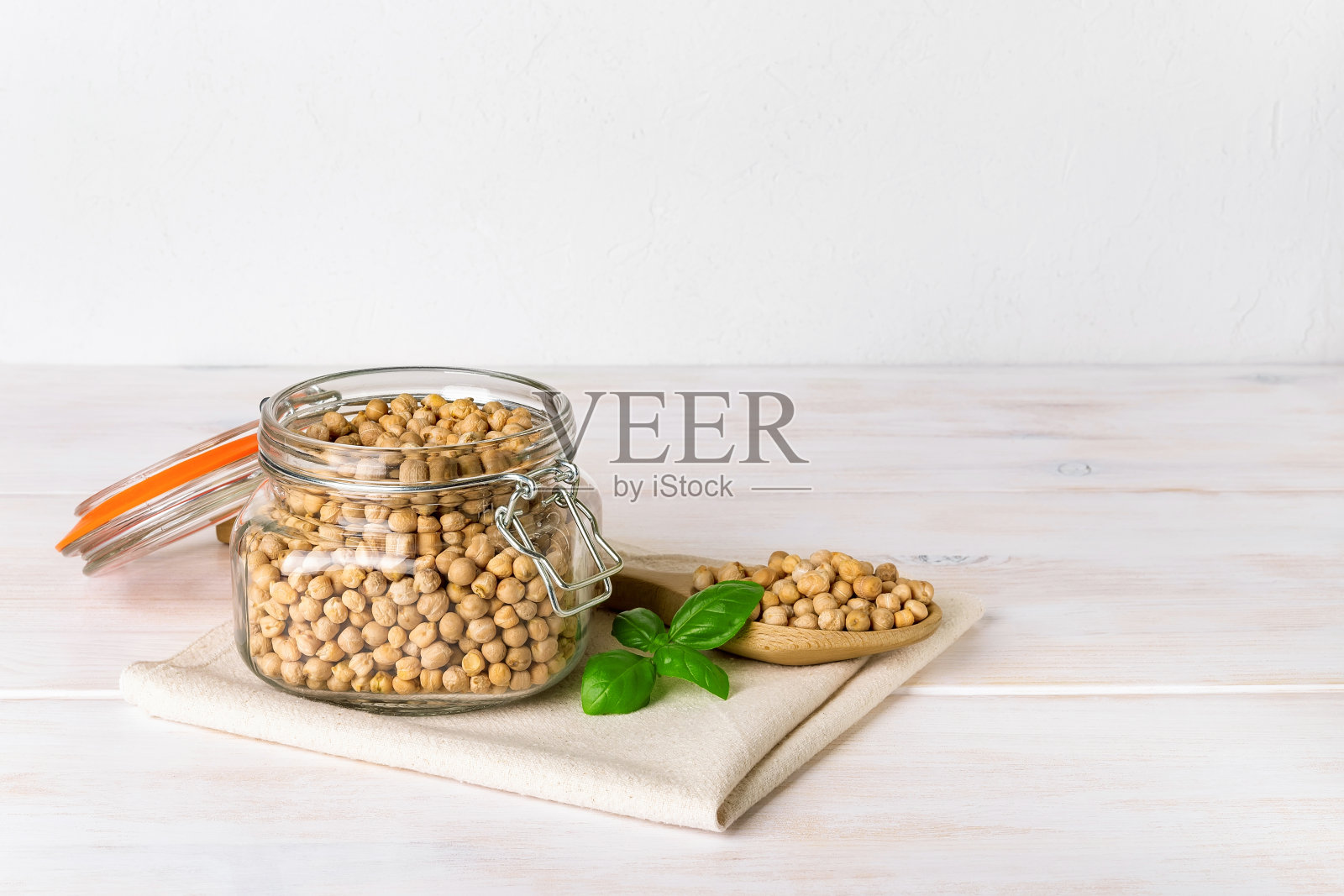 生的鹰嘴豆放在一个夹在顶部的玻璃罐子里，一个大木勺放在一张白色的质朴的桌子上。干鹰嘴豆用来做鹰嘴豆泥。豆类植物蛋白和膳食纤维的健康饮食。照片摄影图片