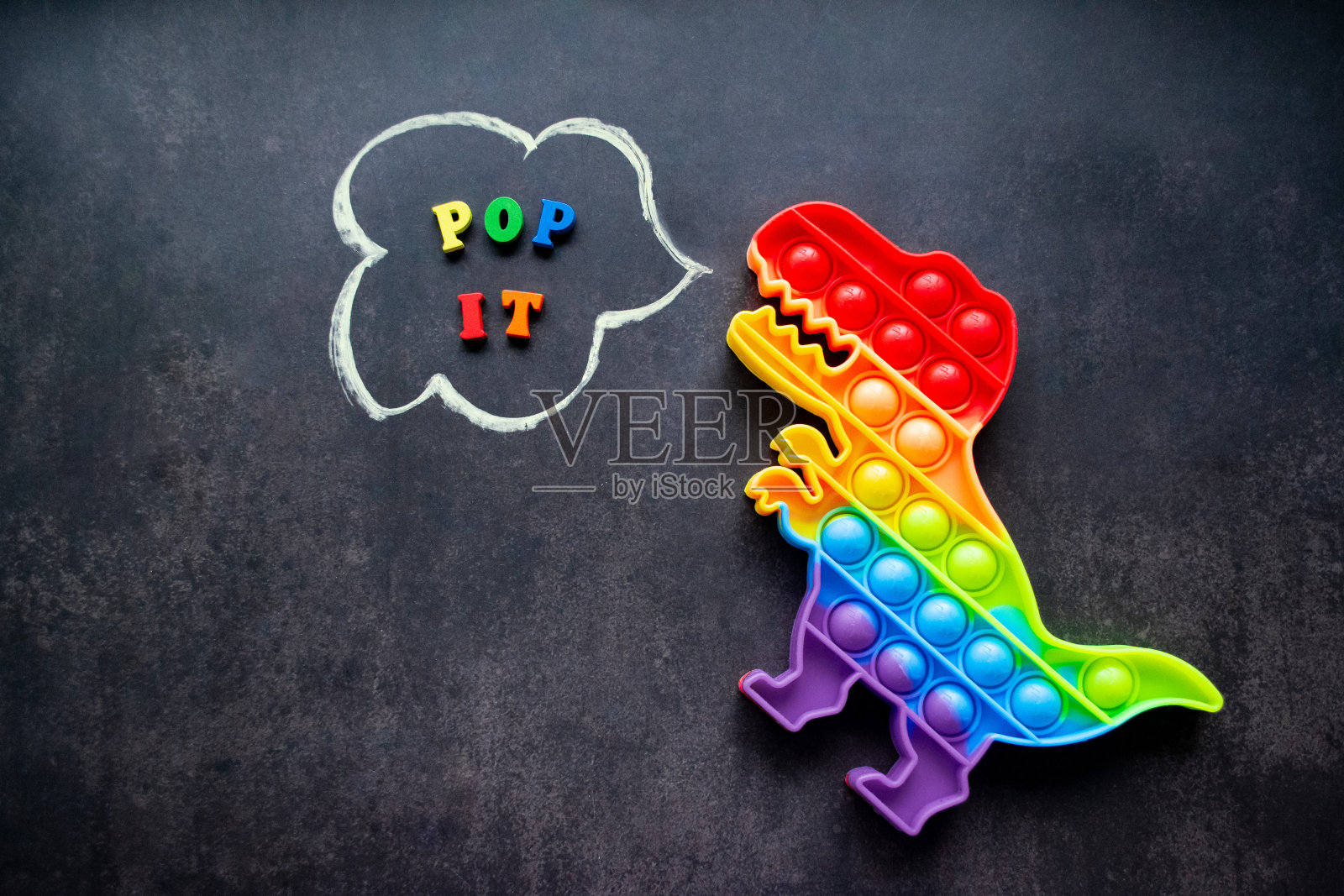 Pop it dinosaur toy rainbow on a black background with五颜六色的字母和题词- Pop it in a speech bubble照片摄影图片