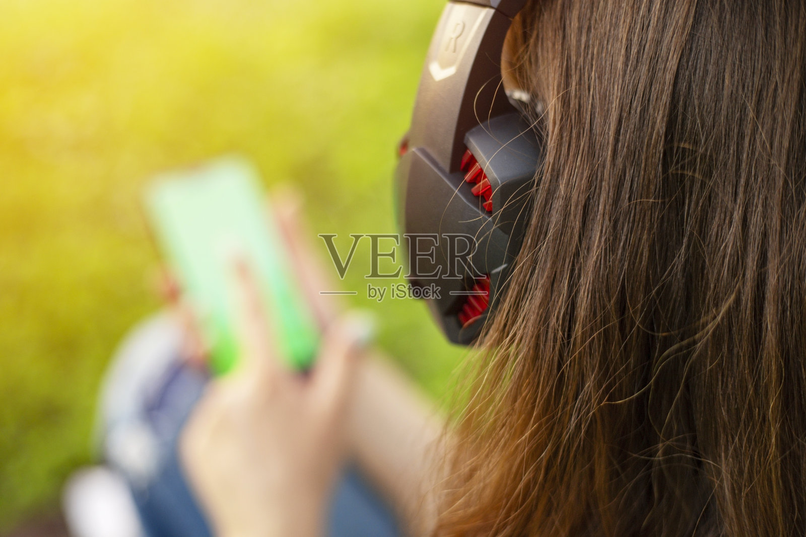 一位女士坐在公园的草地上，手拿智能手机，戴着耳机听音乐。照片摄影图片