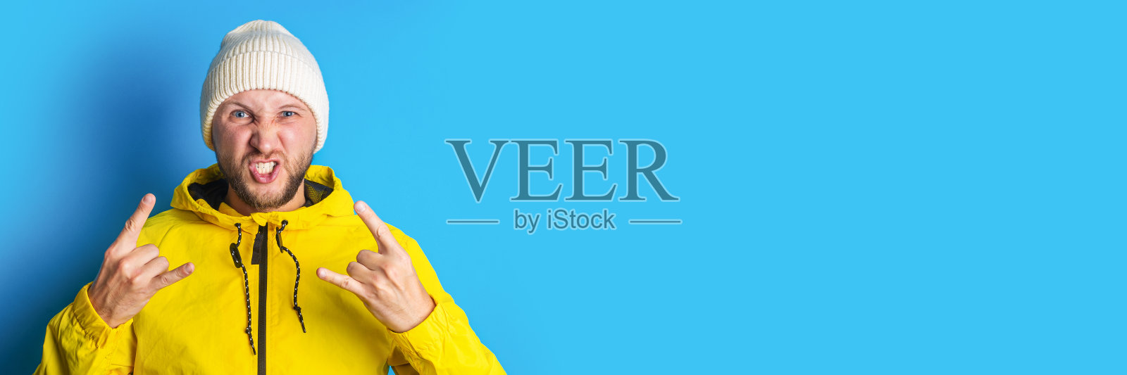 一名身穿黄色夹克、蓄着胡须的年轻人展示了一只摇滚山羊，蓝色背景上有一只角。横幅照片摄影图片