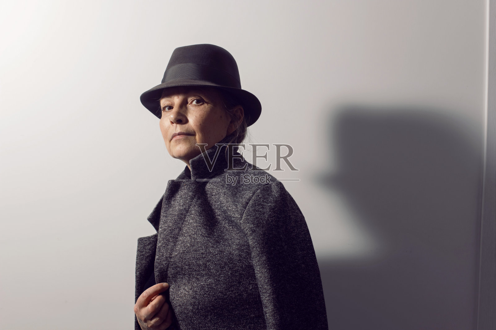 在办公室里，一位穿着黑衣服、戴着帽子的时髦老妇人靠着一堵白墙站着照片摄影图片