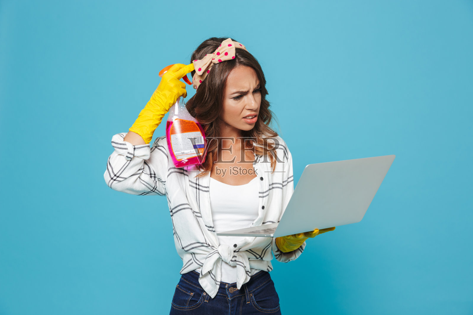 在清洁过程中，戴着黄色橡胶手套，手持清洁剂喷雾器和笔记本电脑的困惑困惑的家庭主妇的肖像，在蓝色背景上隔离照片摄影图片