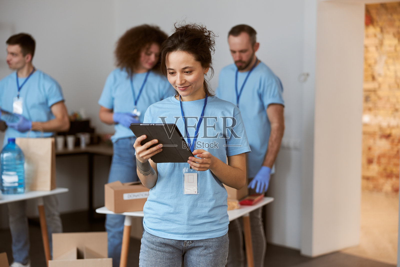 年轻迷人的女志愿者穿着蓝色制服，微笑着使用平板电脑，站在室内。团队整理，打包物品在后台照片摄影图片