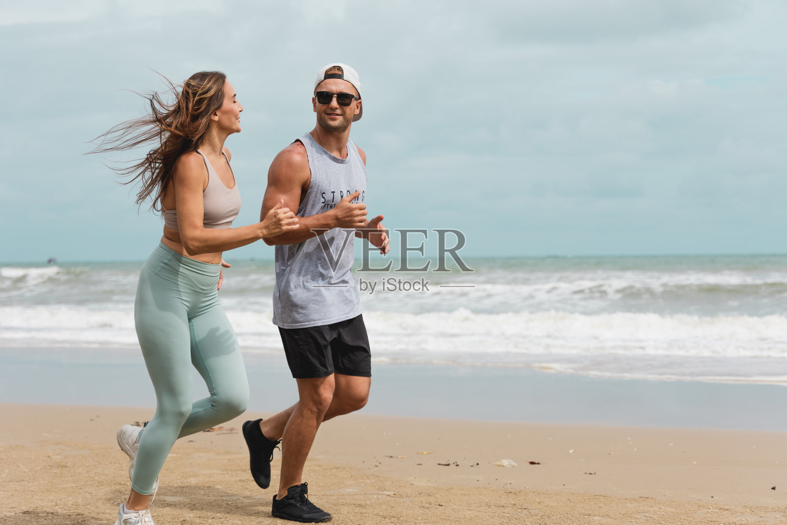 一个穿着运动服的白人男人和女人在海滩上慢跑。年轻的健身运动夫妇戴着耳机在海边跑步。健康运动的概念照片摄影图片
