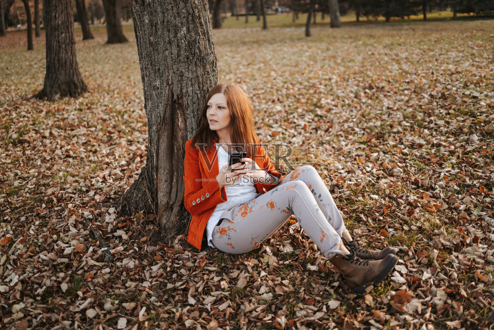 美丽的年轻女子穿着时髦的衣服坐在公园的树下在秋天和使用智能手机照片摄影图片