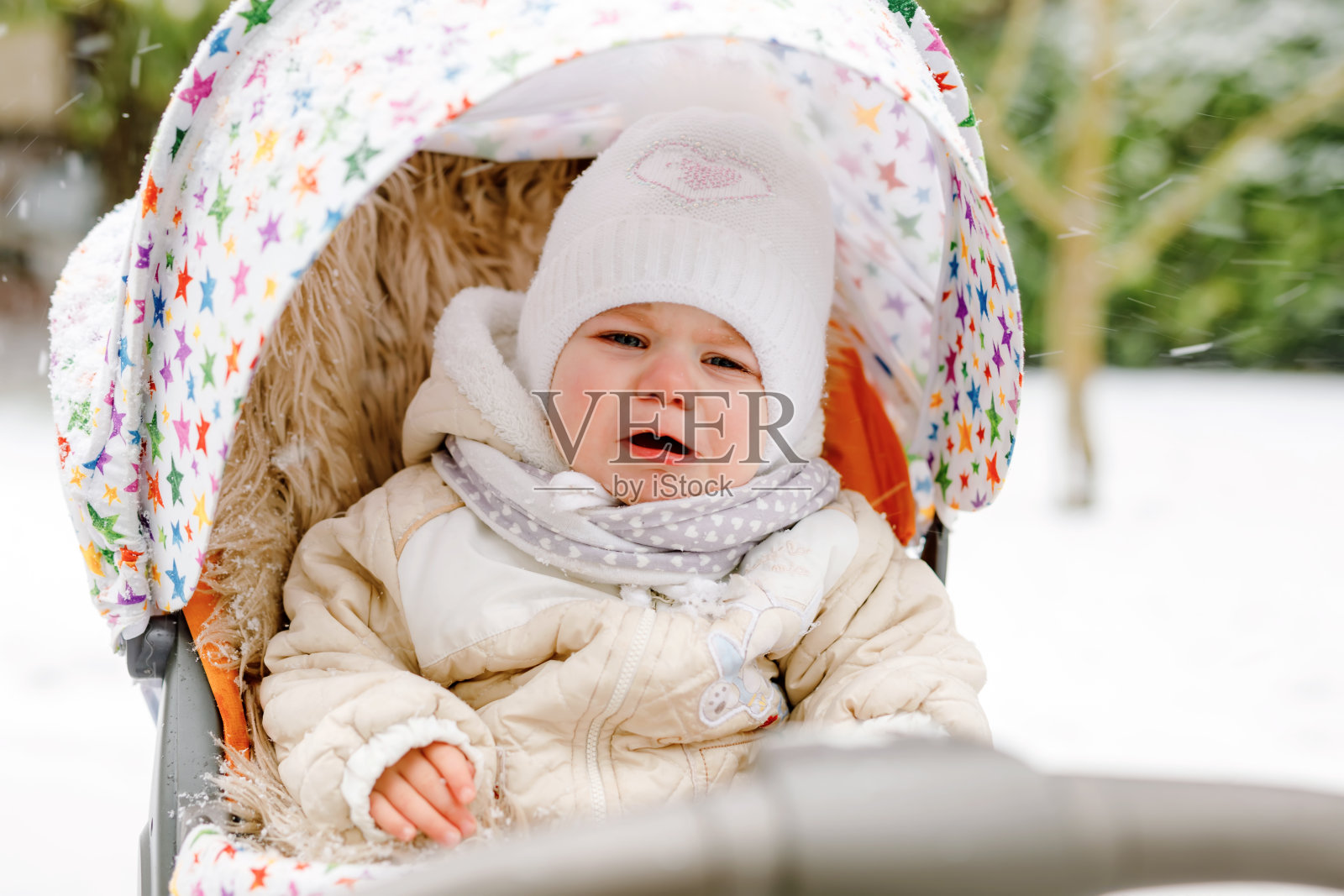 冬天里，哭闹的小女孩坐在婴儿车里。闷闷不乐，心烦意乱，疲惫不堪的孩子穿着暖和的衣服。婴儿的第一场雪。冬季在户外散步。照片摄影图片