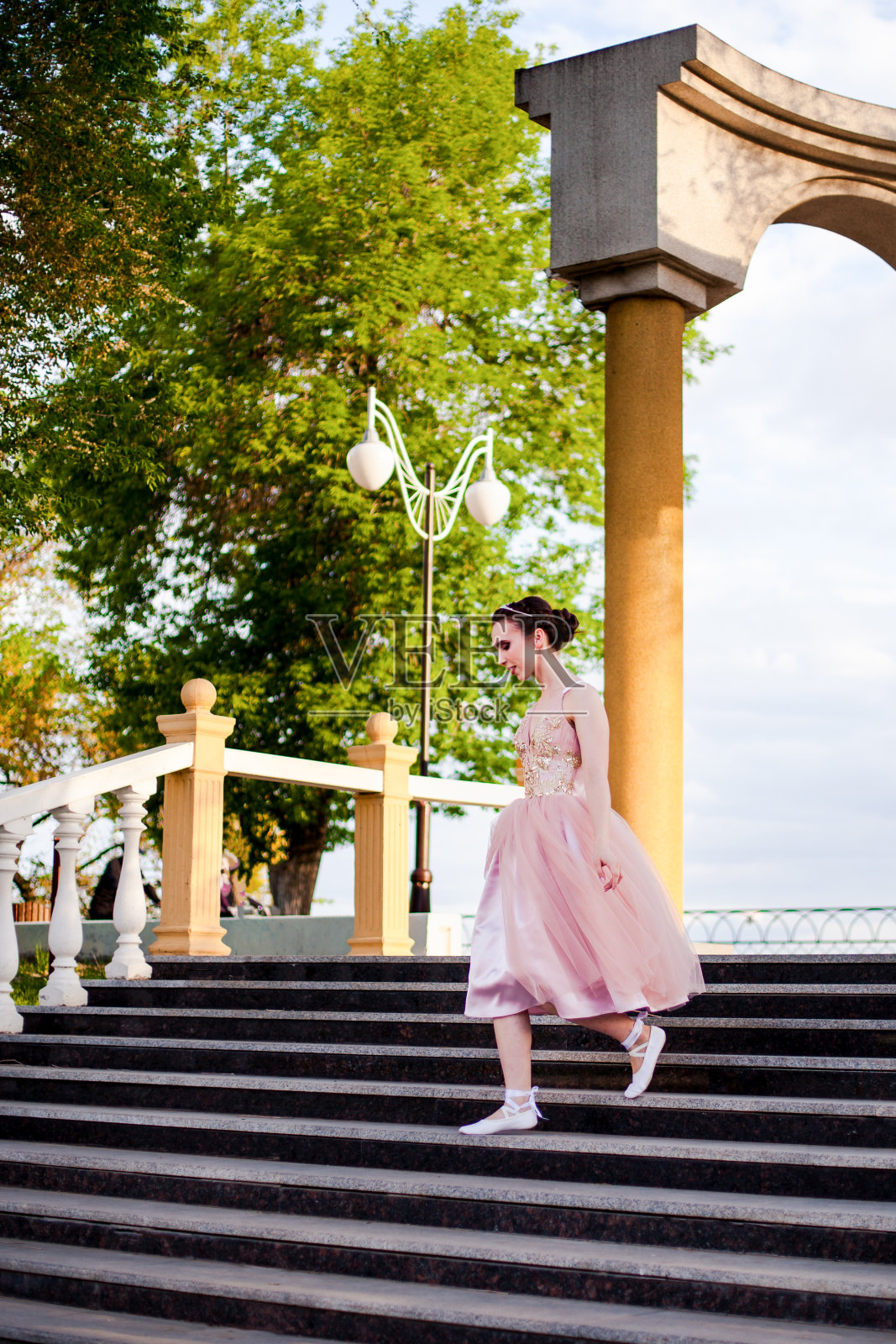 夏天，在城市公园的河岸上，一个年轻的芭蕾舞女演员穿着粉红色的丝绸连衣裙优雅地跑下有柱子的拱形台阶照片摄影图片