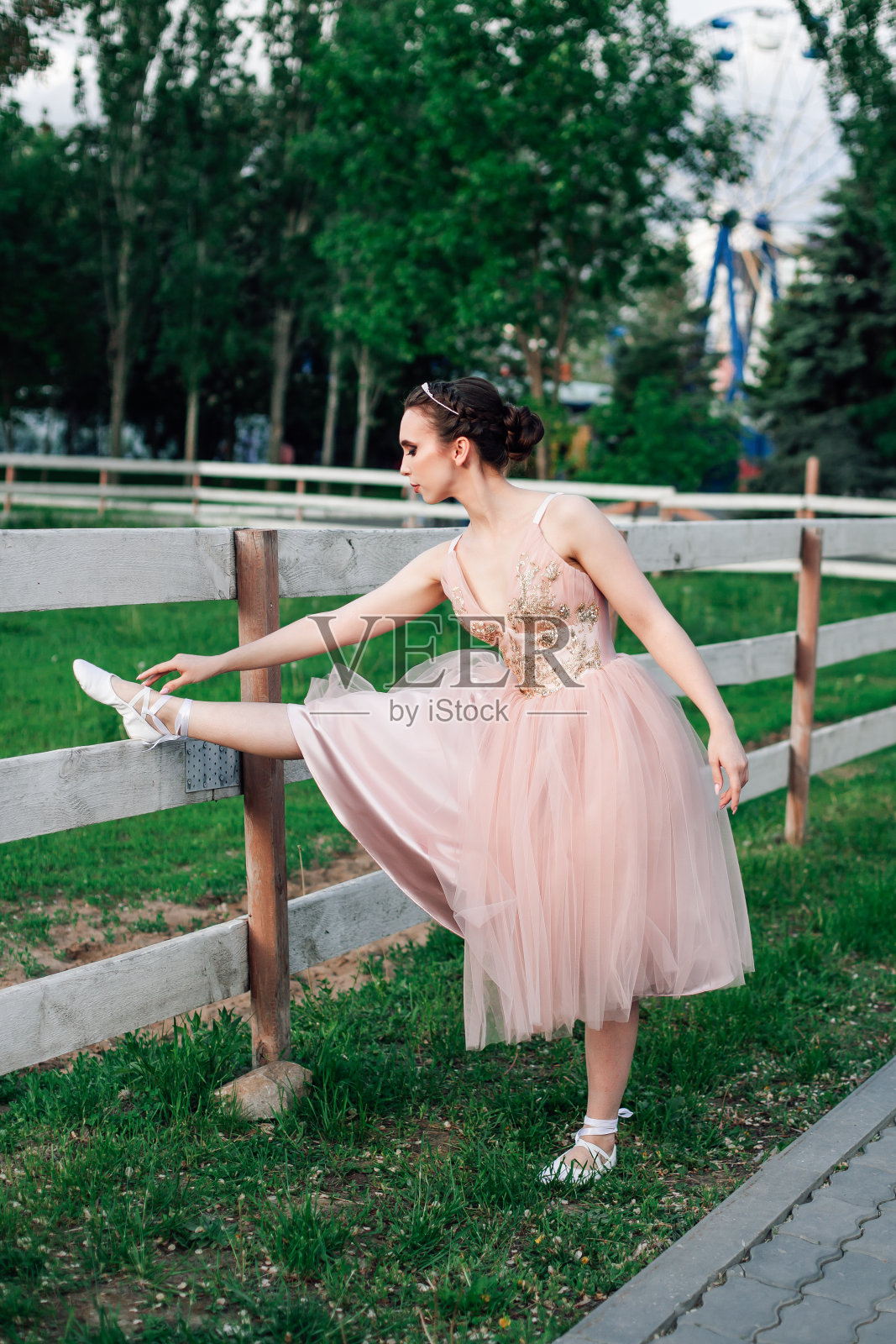 一个年轻的芭蕾舞女演员穿着粉红色的裙子在一个有绿色草地和树木的公园里做伸展运动在夏季日落照片摄影图片