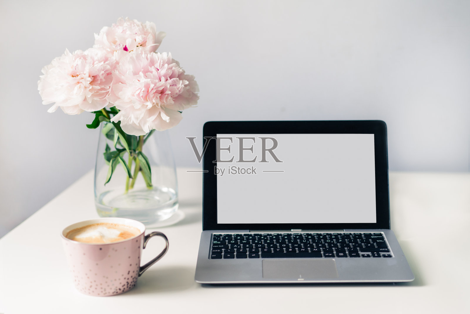 舒适的工作场所。白色的办公桌上放着一杯新鲜的咖啡，打开的笔记本电脑上有空白屏幕，花瓶上插着盛开的牡丹花。极简主义的工作区。自由职业，在线学习，家庭办公。照片摄影图片