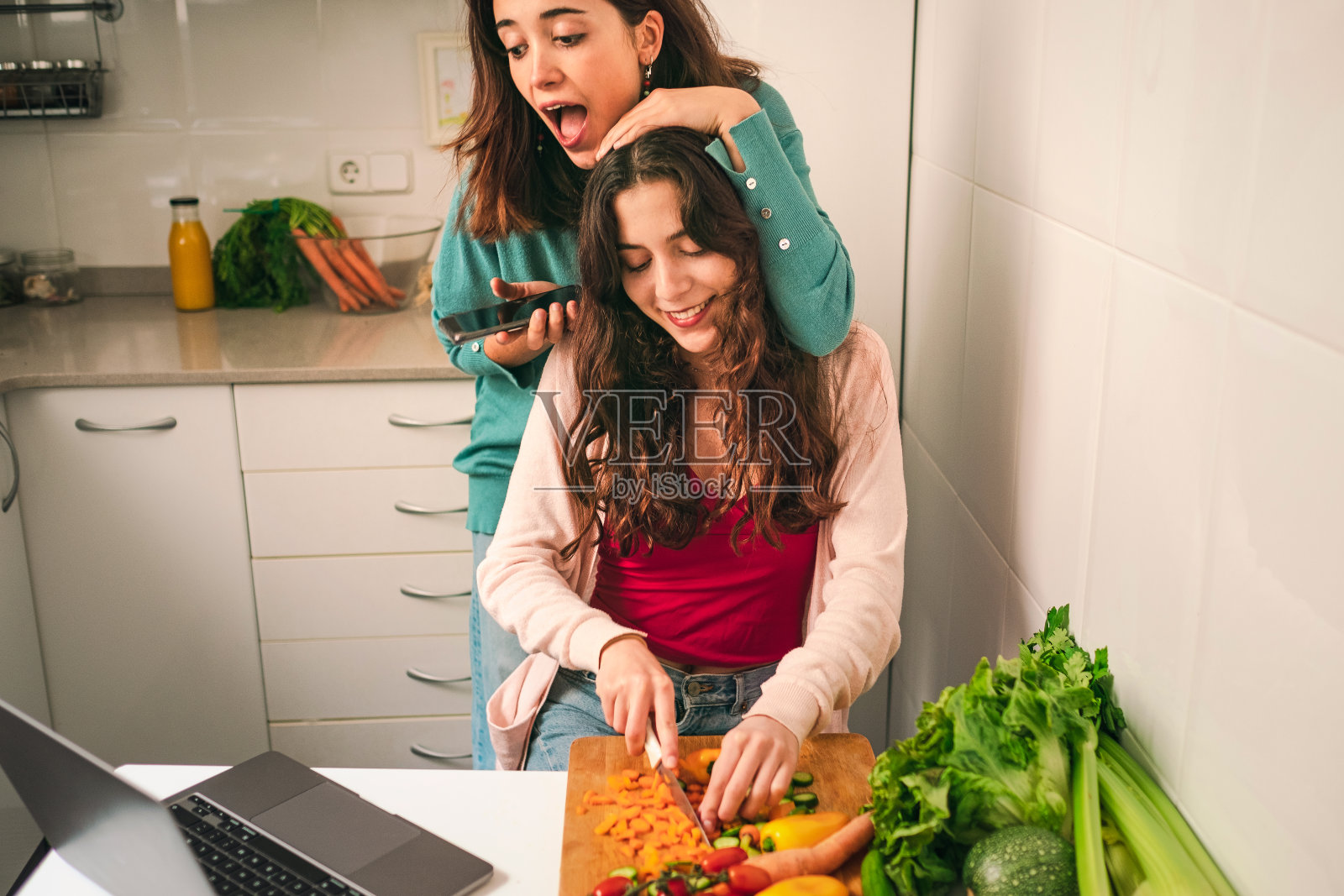 惊讶的少女在笔记本电脑上查看社交媒体，同时她拥抱了她准备健康餐的姐姐照片摄影图片