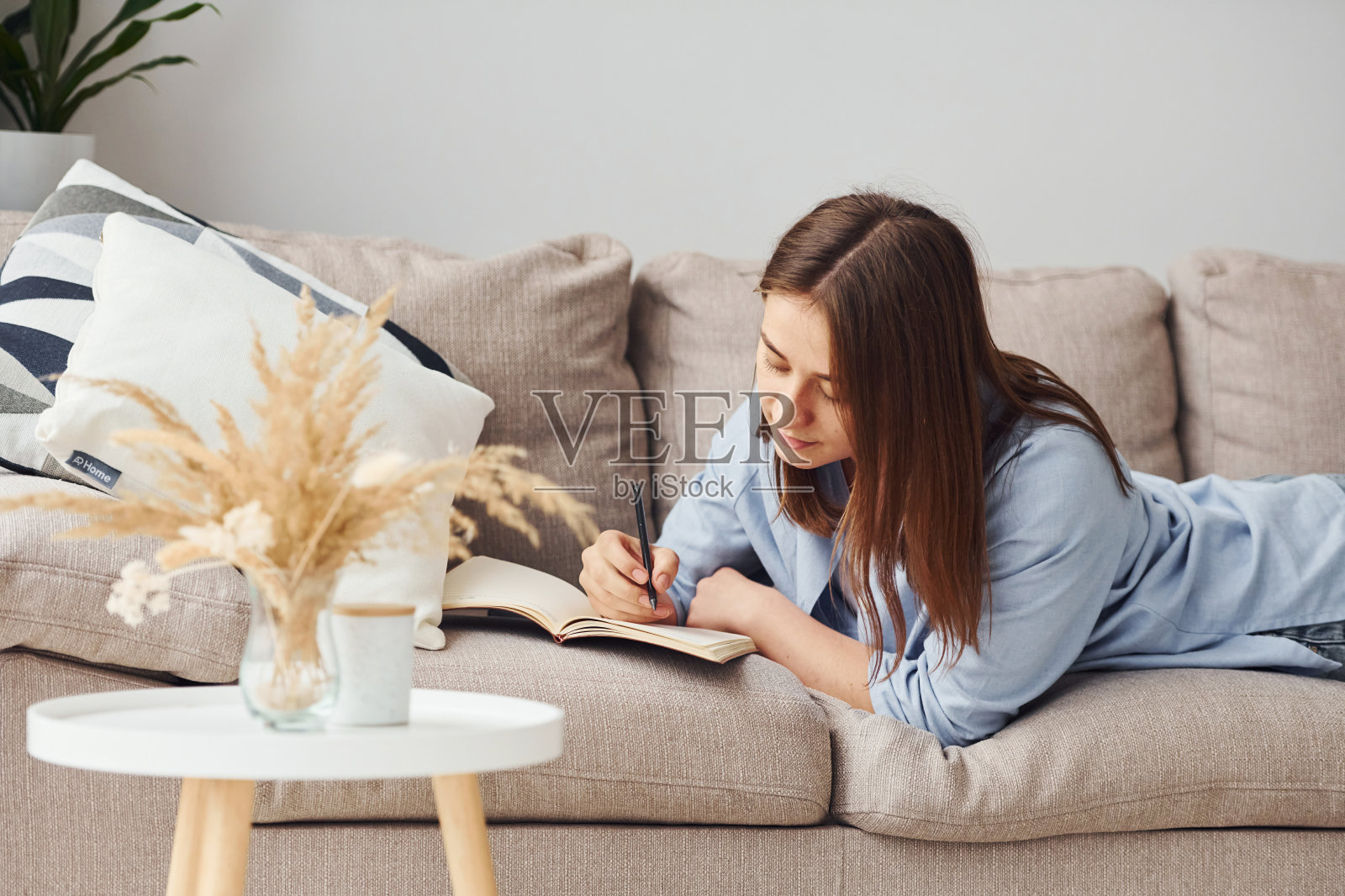 穿着牛仔裤和蓝色衬衫的漂亮年轻女子躺在沙发上，拿着笔记本和笔照片摄影图片