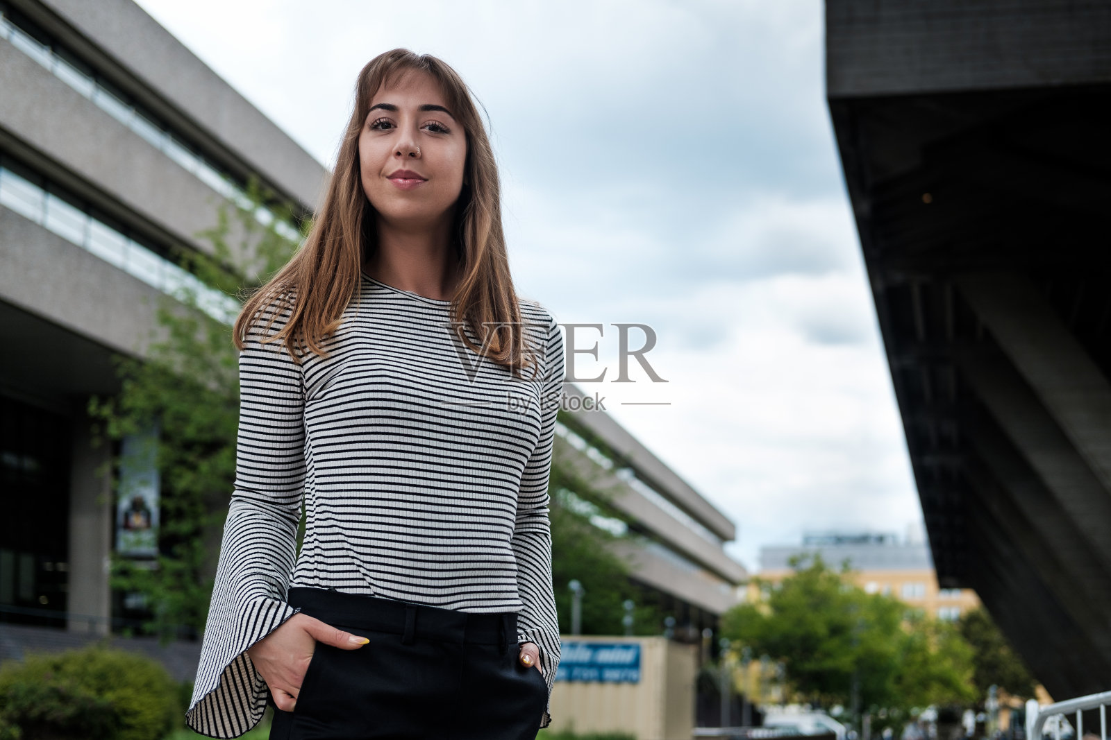 年轻自然的女人摆姿势看着相机与她身后的建筑。照片摄影图片