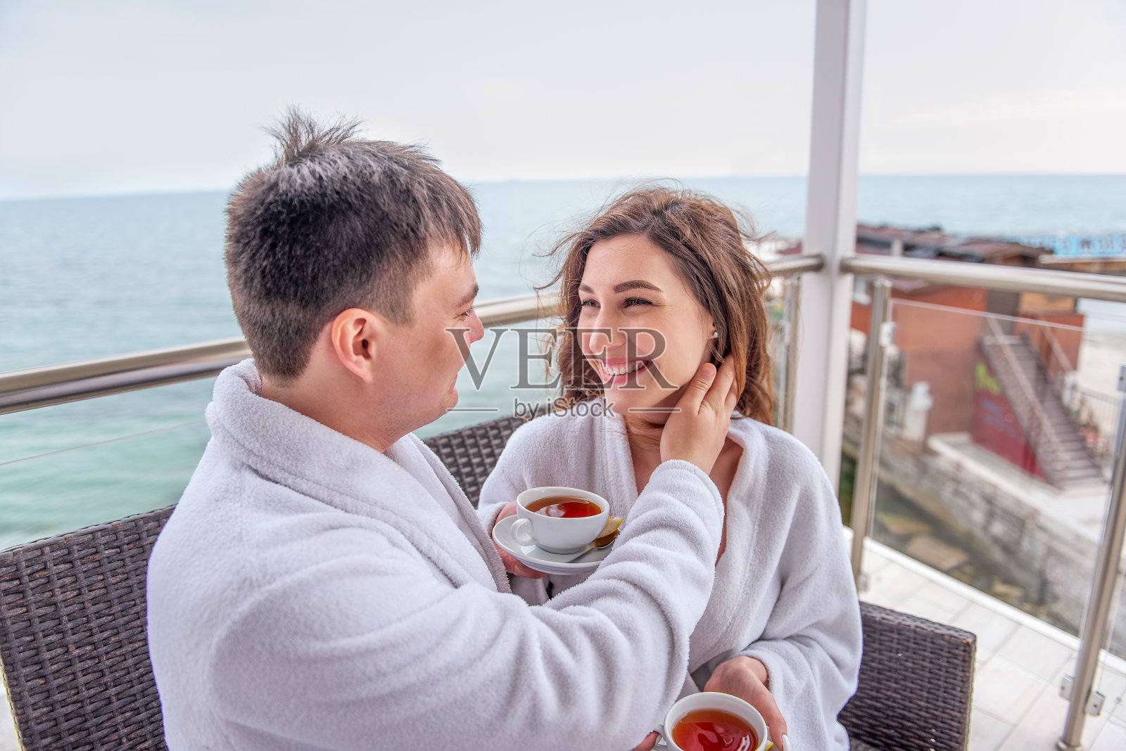 一对穿着白色浴袍的夫妇坐在海边的露天露台上，用杯子喝柠檬茶。情人度假旅行。一个年轻男子为女人拉直头发。新婚夫妇度蜜月的地方照片摄影图片