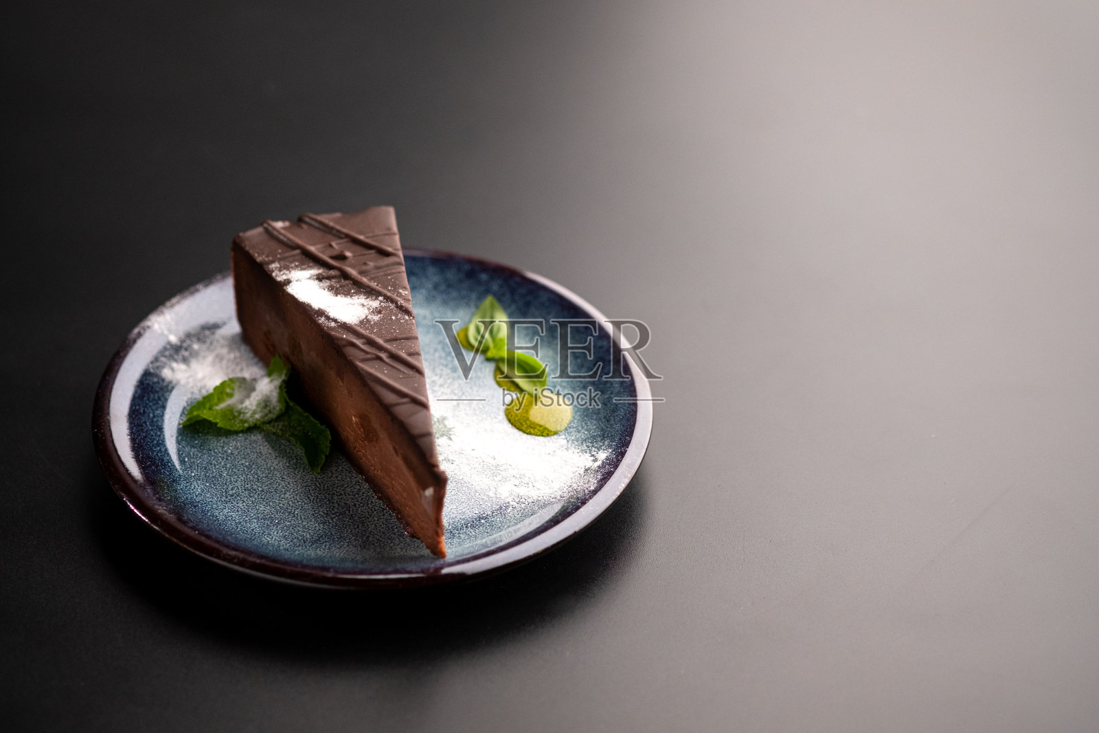 一块巧克力芝士蛋糕放在一个蓝色的盘子里，用糖粉和薄荷叶装饰。黑色背景。照片摄影图片