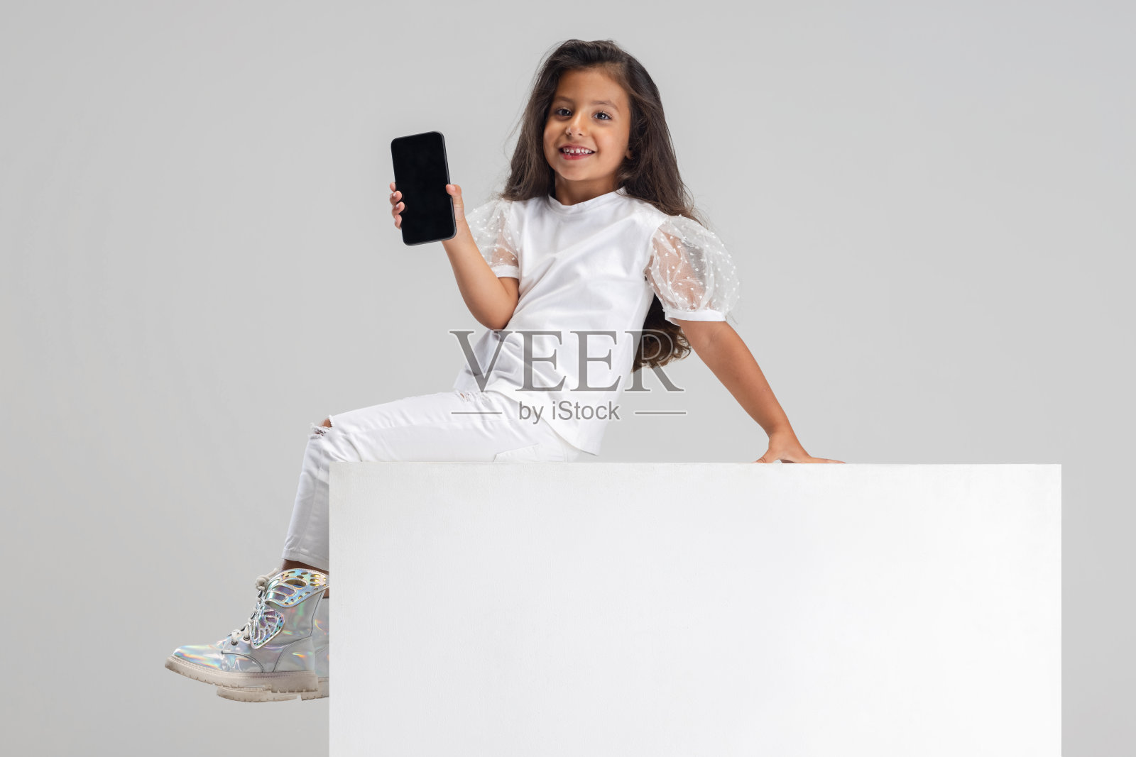 小可爱微笑的白人女孩坐在大盒子上的肖像与智能手机隔离在白色的工作室背景。照片摄影图片