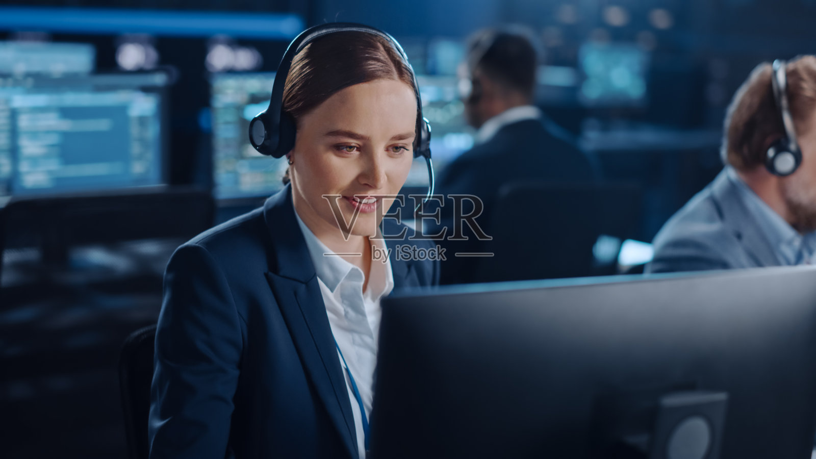 在一个黑暗的监控室里，满是同事和显示屏，快乐美丽的技术客户支持专家一边用耳机说话，一边用电脑工作。照片摄影图片