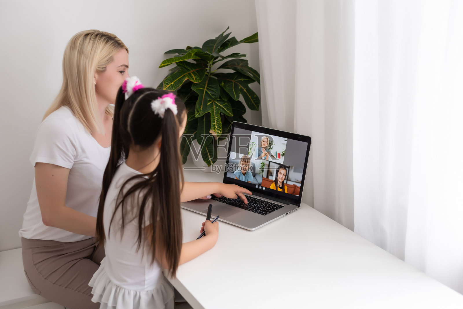 由于冠状病毒大流行，家庭学校的小女孩通过数字远程互联网会议从学校老师那里学习在线课程。孩子看着电脑，在妈妈的帮助下写纸条照片摄影图片