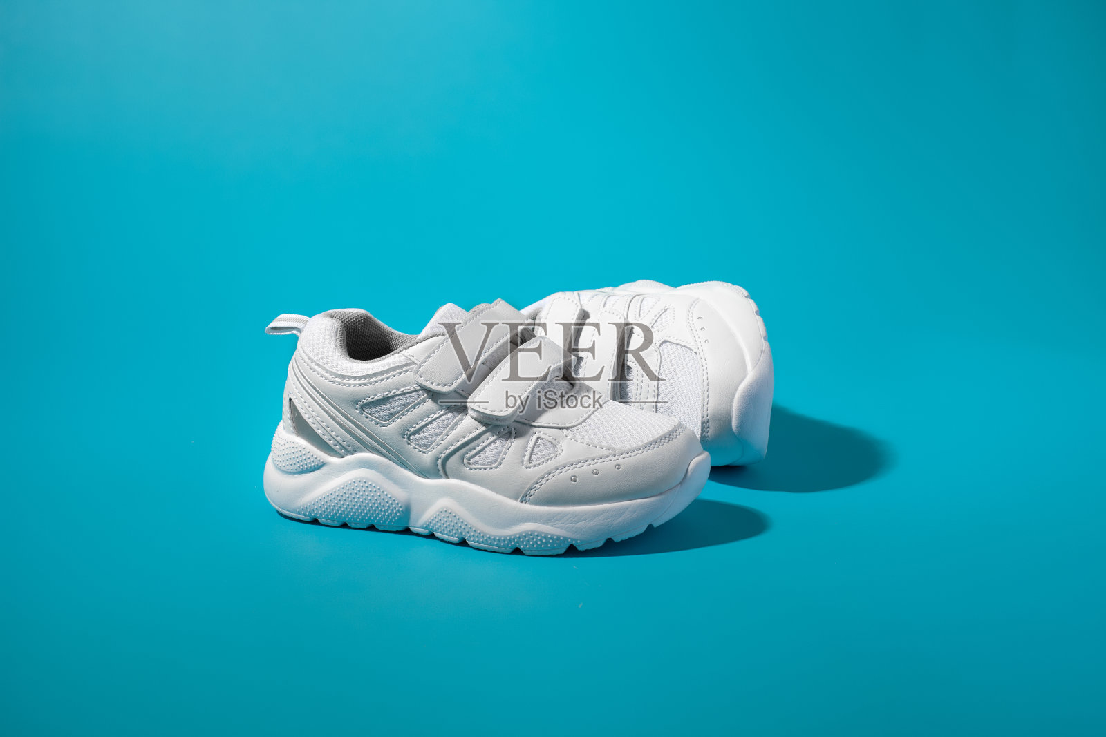 一只带尼龙搭扣的白色儿童跑鞋放在另一只的前面，另一只则侧卧在蓝色的纸背景上，有坚硬的阴影照片摄影图片