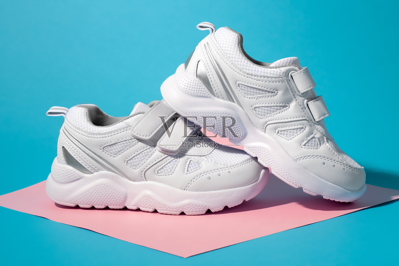 特写侧视图的两个白色青少年运动鞋与尼龙搭扣扣在一个几何方形纸粉红色和蓝色的背景与硬阴影，一个运动鞋站在另一个照片摄影图片