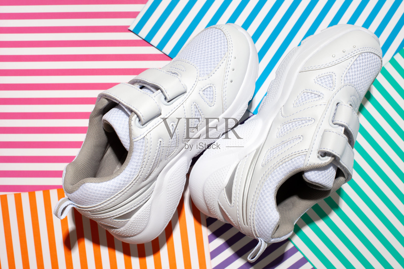 俯视图两白色男女皆可穿的运动鞋与尼龙搭扣，便于鞋上的抽象几何纸上的彩色条纹背景与硬光照片摄影图片