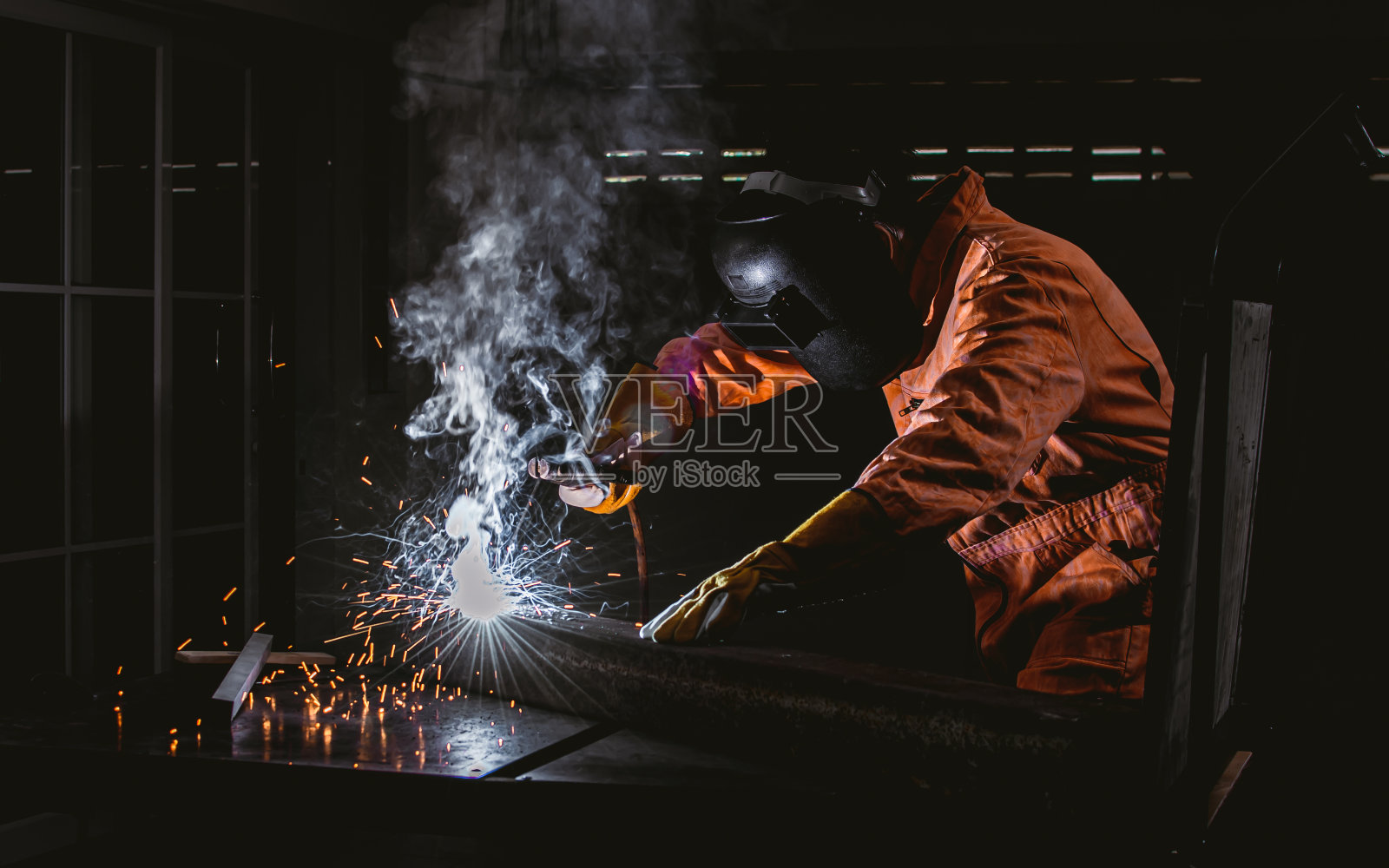 亚洲英俊的胡子男机械师穿着橙色制服和安全面具，使用设备和焊接铁或金属有意向在工业工厂。照片摄影图片