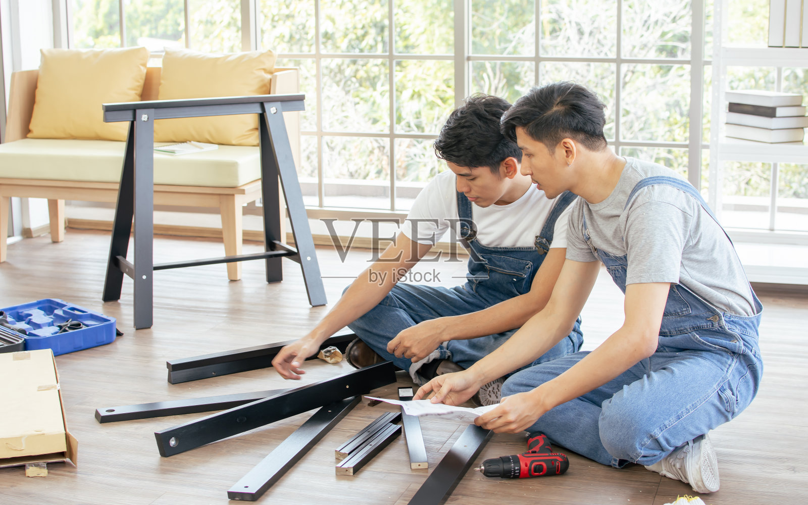 两个身穿蓝色工装裤的亚洲帅哥，在家里按照手工操作互相帮助或支持组装DIY家具。照片摄影图片