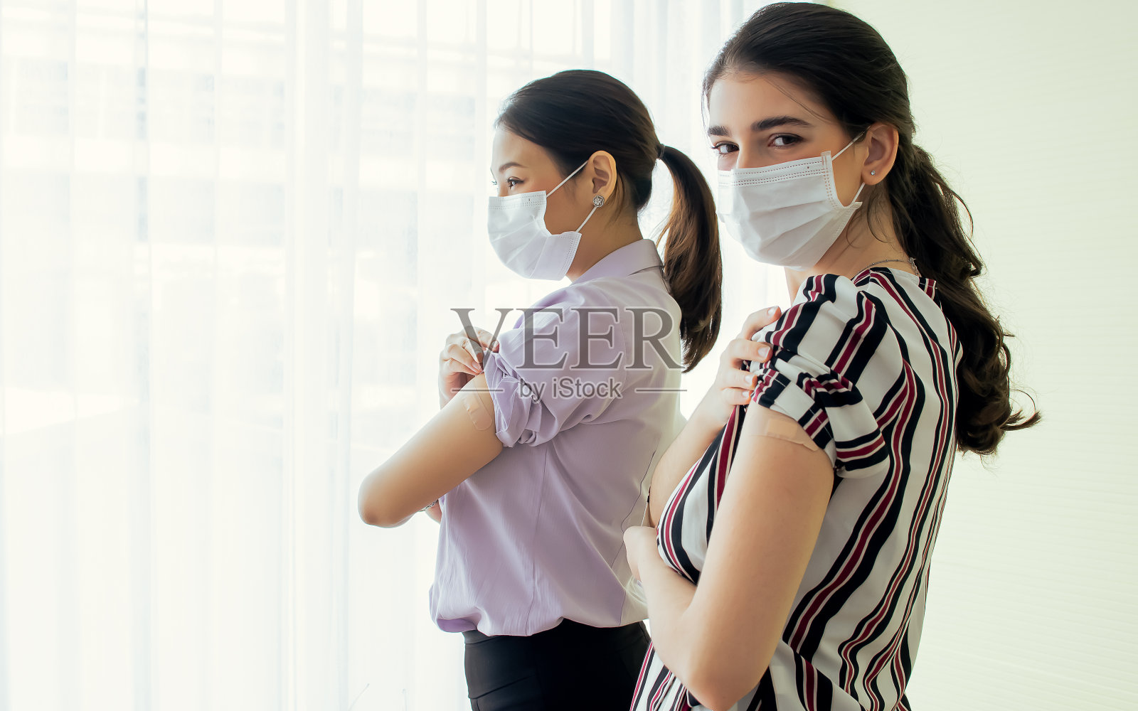 描绘两名亚洲和高加索妇女戴着口罩，展示绷带，接受免疫疫苗保护病毒，信心十足地看着镜头。医疗和大流行危机概念。照片摄影图片