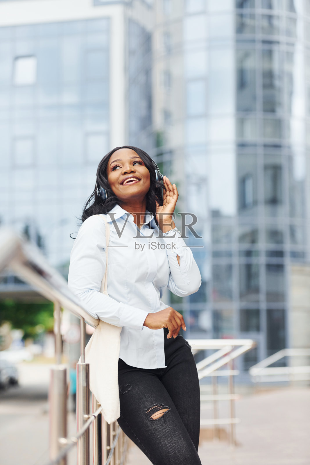 戴着耳机听音乐。穿着白衬衫的年轻美国黑人妇女在城市的商业大厦外照片摄影图片